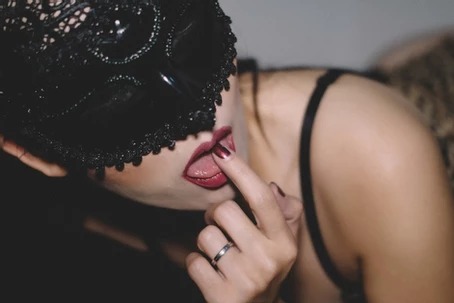 A woman in a black mask and red lipstick biting her finger.