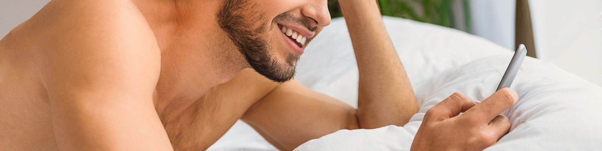 A shirtless person laying down on a bed with white sheets smiling while looking at a phone.